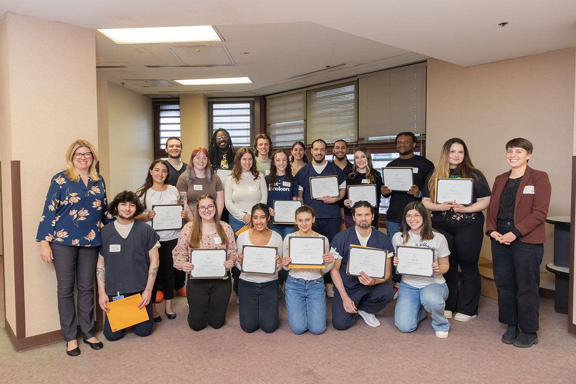 A group of about 20 smiling college students and students who are incarcerated hold up completion certificates